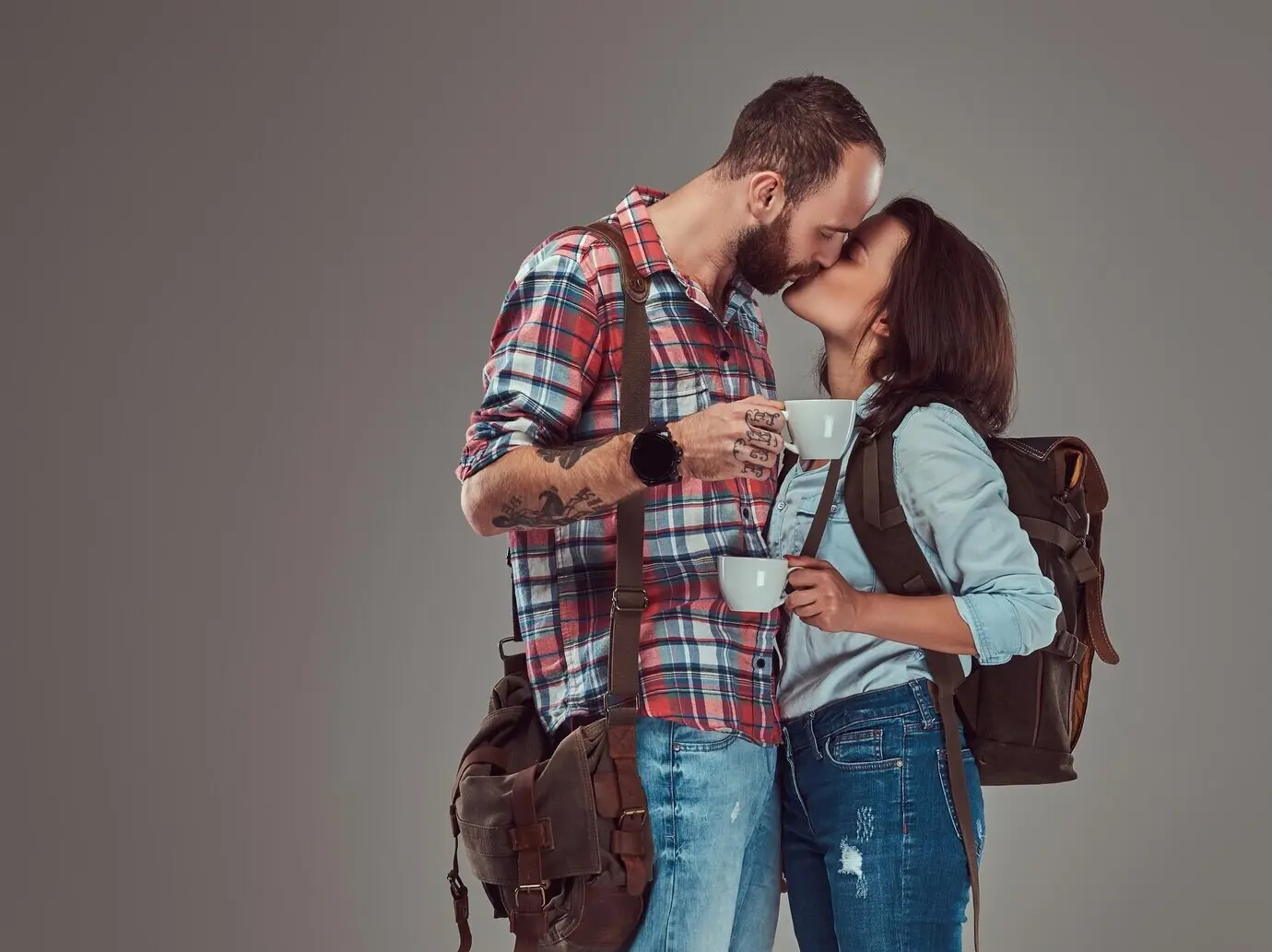 A happy tourist couple kisses while holding cups of coffee, isolated on a gray background.