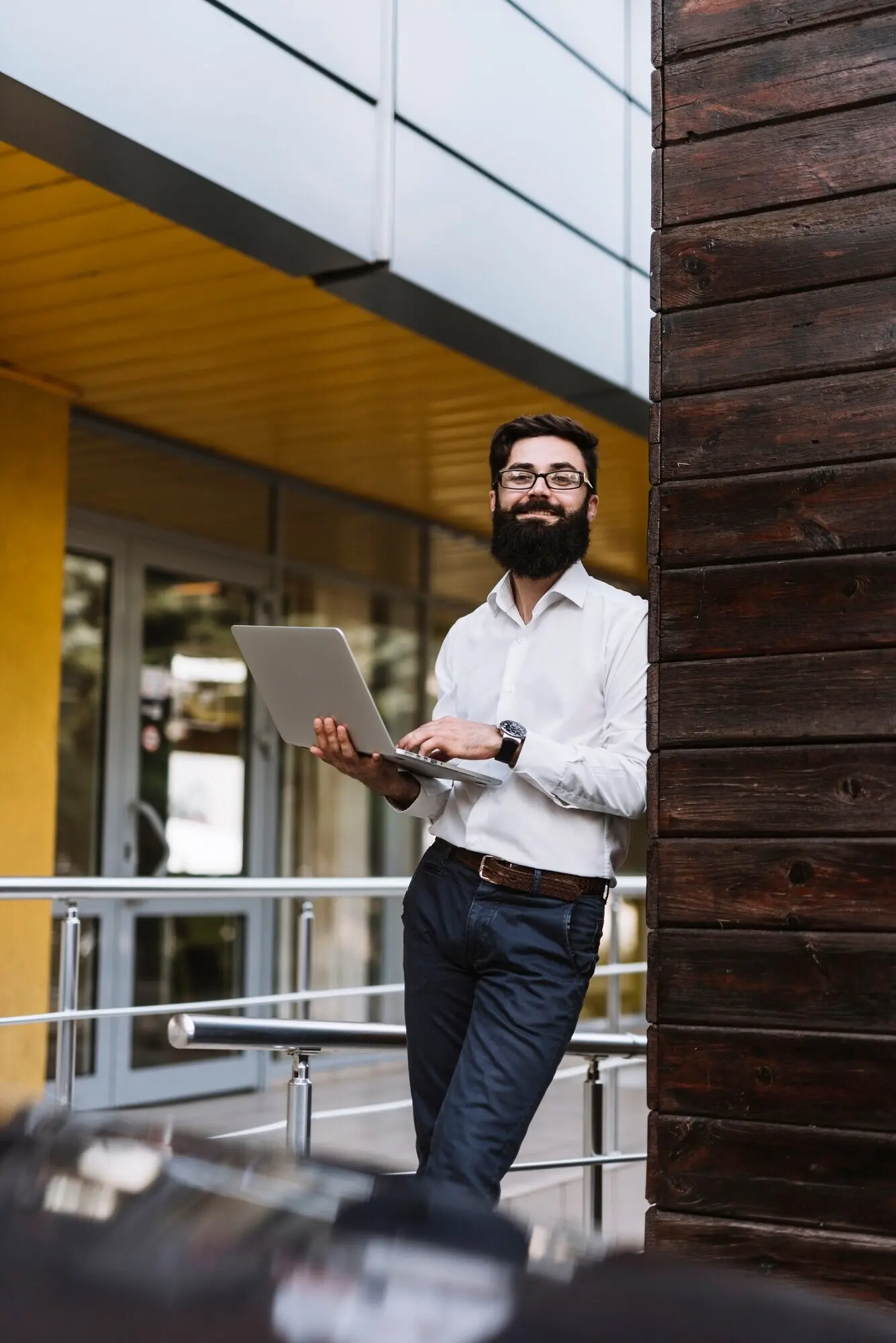 A smiling young businessman leans against a wall while holding a laptop.