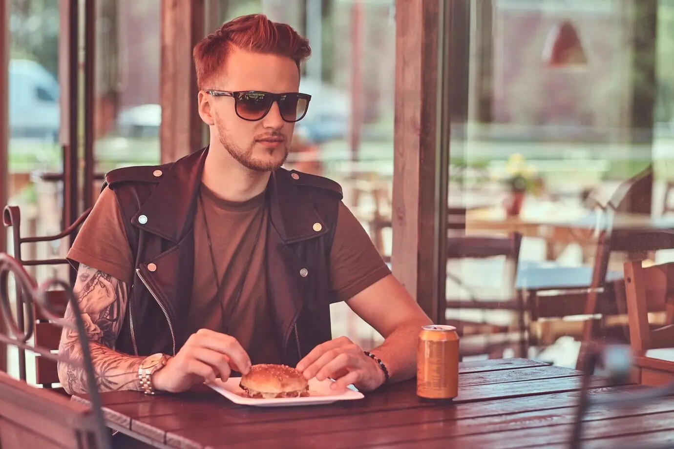Portrait of a handsome, stylish hipster guy seated at a table, dining at a roadside cafe with a hamburger and a soda he ordered.